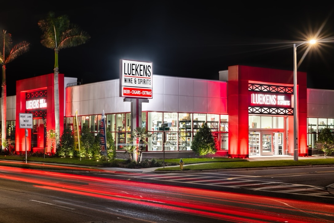Night view of a brightly lit Luekens Wine & Spirits store, with red exterior lighting and light trails from passing cars on the road in front.