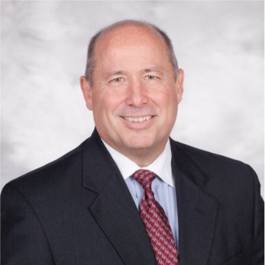 A man in a suit and red tie smiling against a gray background.