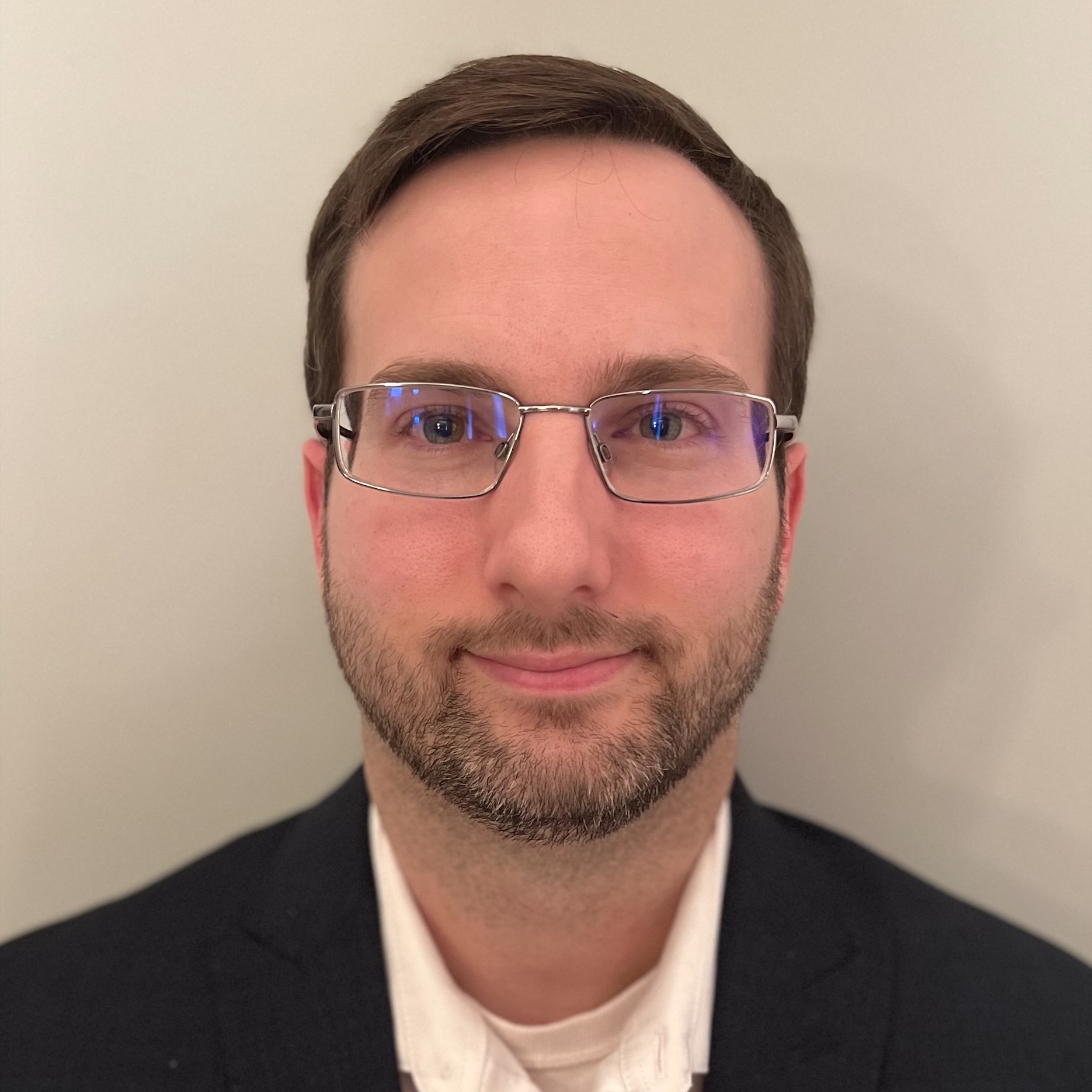A man with glasses, short brown hair, and a trimmed beard is wearing a dark suit jacket and white shirt, facing forward against a plain background.