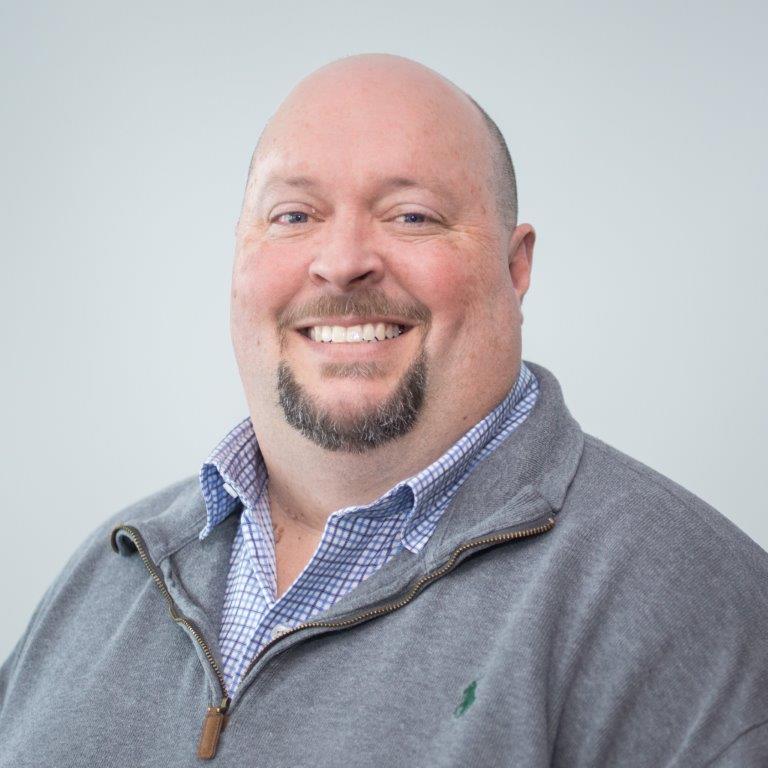 A man with a goatee, wearing a gray quarter-zip sweater over a checkered shirt, smiles while facing the camera.