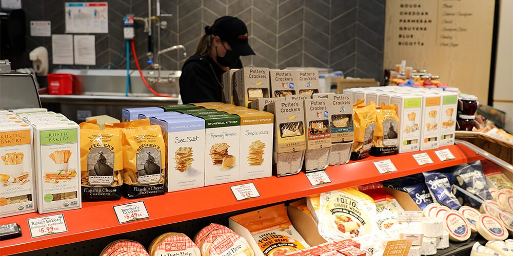 A dairy counter with cheeses in the front. In the background, a grocery store attendant is looking down
