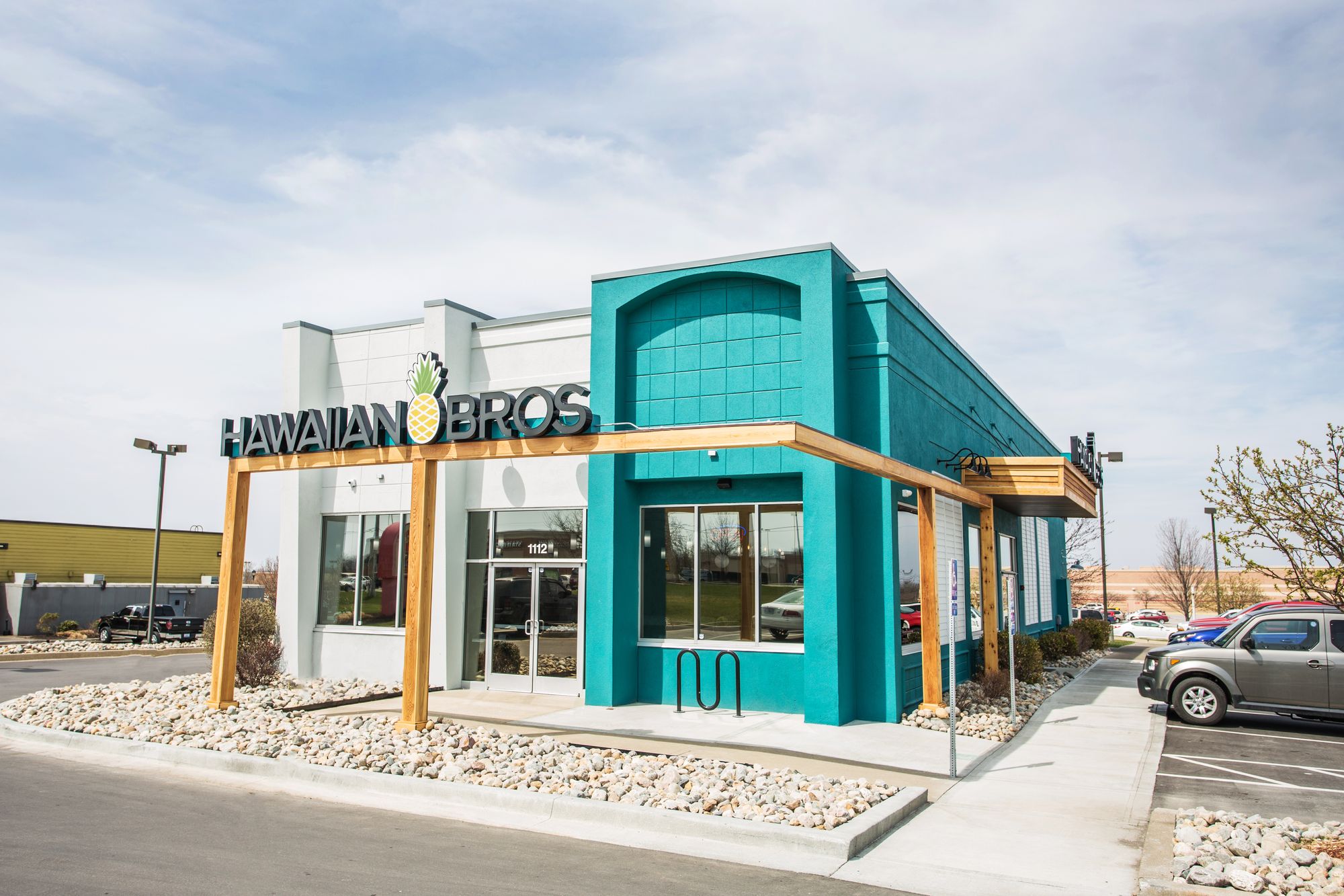 A Hawaiian Bros restaurant with a turquoise facade, large windows, and a wooden pergola at the entrance. Cars are parked to the right. The sky is overcast.