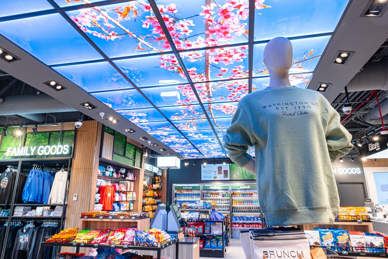 A retail store interior features snacks, drinks, and souvenirs, with a mannequin in a sweatshirt and a ceiling display of cherry blossom imagery overhead.