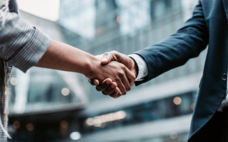 Two people shaking hands in an outdoor urban setting, with buildings visible in the background.