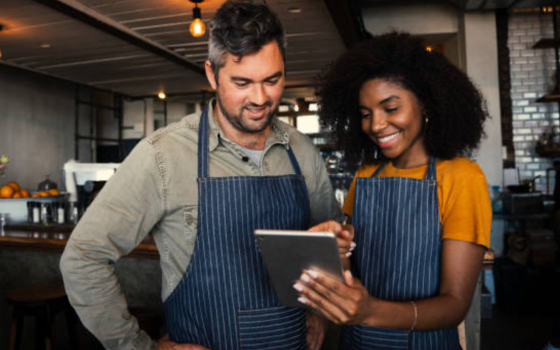 Two people wearing aprons stand in a cafe, looking at a tablet and smiling.