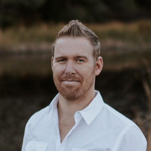 Man with short hair and beard wearing a white shirt stands outdoors with blurred background.