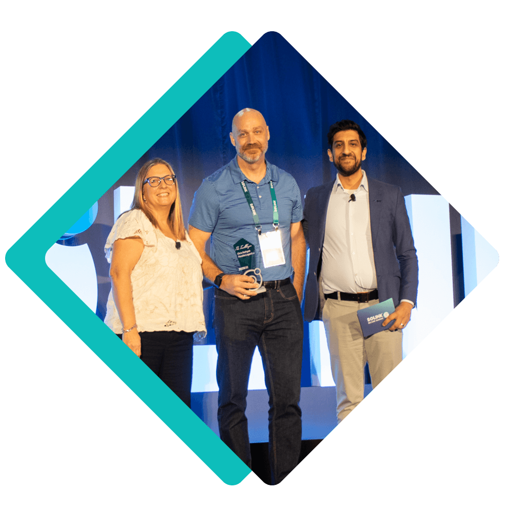 Three people stand on a stage, one holding an award trophy and wearing a lanyard, all smiling for a photo. Blue backdrop and geometric frame shapes accent the image.