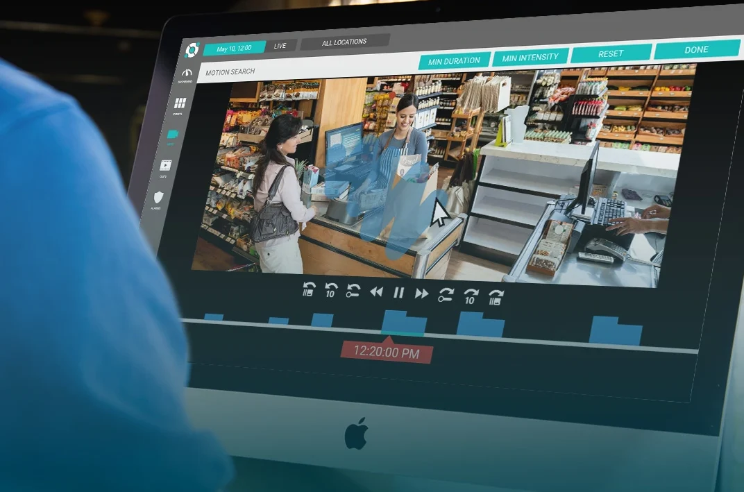 A person in front of an iMac monitor reviewing security footage of a grocery store checkout. The screen shows a customer and cashier interacting.
