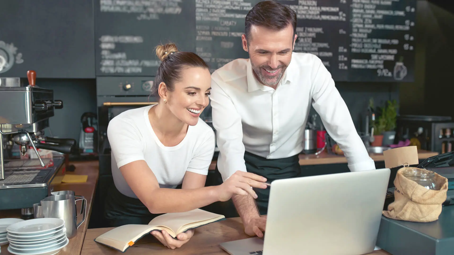 Two people in a cafe look at a laptop screen. One holds a notebook. Chalkboard menu and coffee equipment are in the background.